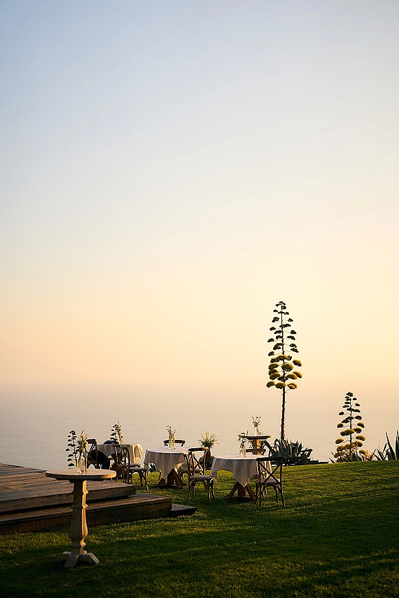 Outdoor reception setup with round tables in white linens, wooden chairs, and bud vase florals on a lawn overlooking the ocean at sunset