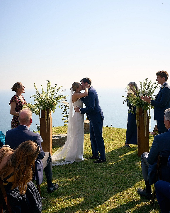 Ceremony kiss as bride in strapless dress and veil kisses groom in navy suit at an outdoor wedding ceremony with ocean-view cliffside backdrop