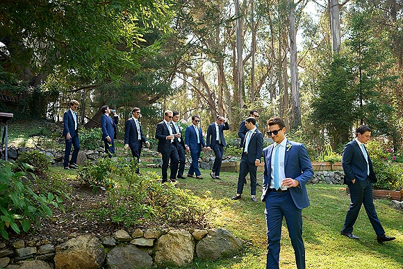Groomsmen portraits of men in navy suits with light blue ties, wearing sunglasses and holding drinks on a garden lawn by stone steps