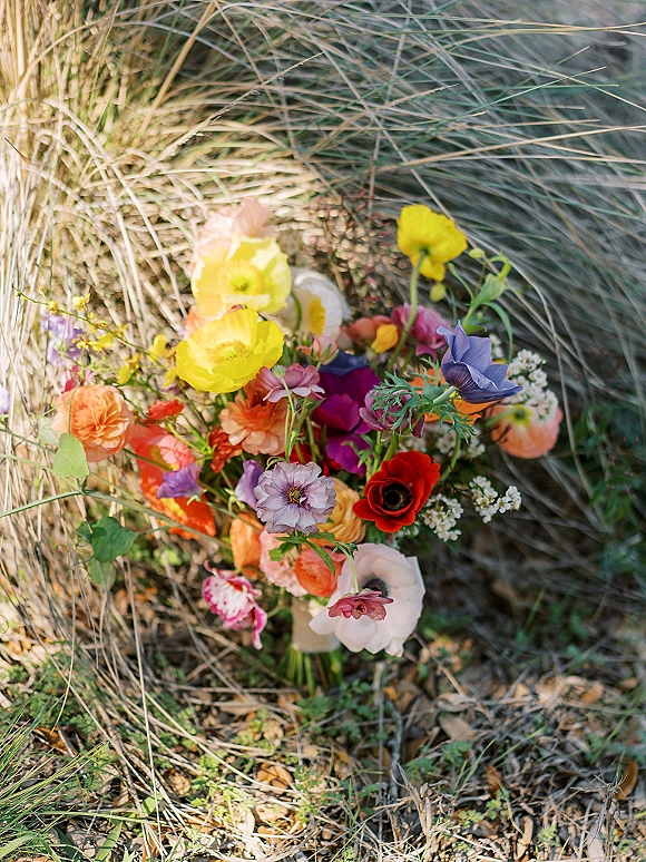Wedding bouquet with wildflower wedding bouquet blooms, greenery, and ribbon resting in tall grass with ground foliage and dried leaves