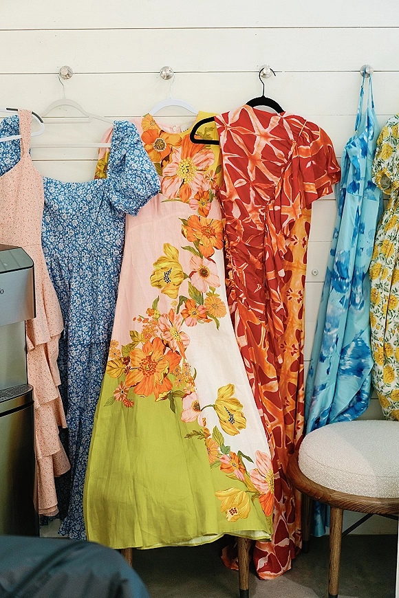 Bridesmaid dresses in mismatched bridesmaid dresses styles hang on wall hooks, a colorful patterned lineup against a white shiplap wall