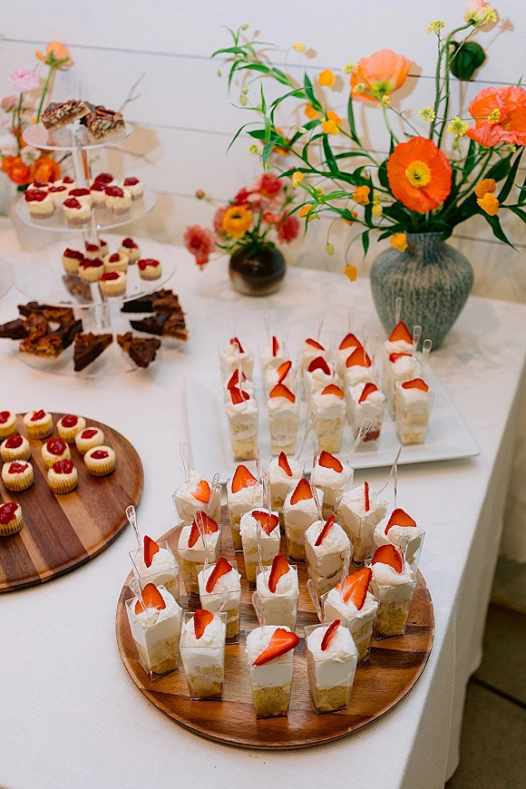 Wedding dessert table with mini dessert cups, strawberry shortcake cups, brownies and cheesecakes on tiered stands with orange florals on white linens