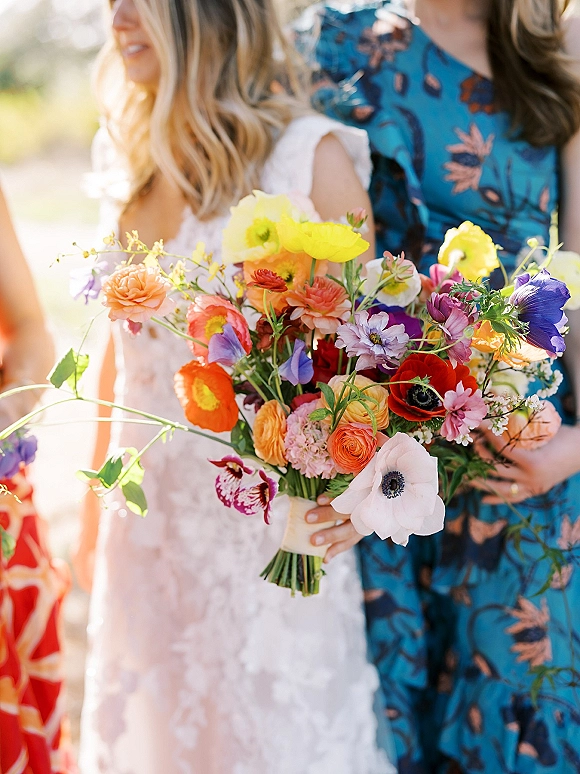 Bridal bouquet of colorful bridal bouquet wildflowers wrapped with ribbon, held against a lace wedding dress in sunlit greenery bokeh background