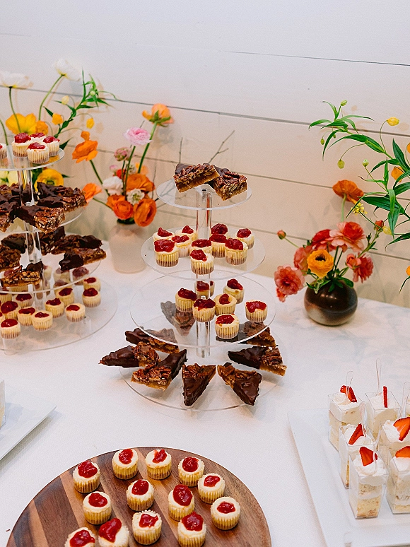 Wedding dessert table with tiered dessert stands holding mini cheesecakes, chocolate pecan bars, strawberry cups, and bud vase florals on white shiplap