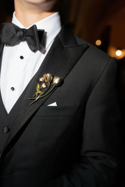 Groom boutonniere, metallic boutonniere pinned to a black tuxedo satin lapel with bow tie and pocket square against warm bokeh lights