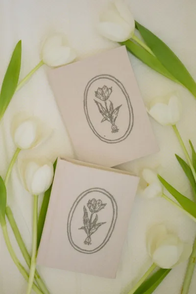 Vow books in a his and hers vow books set, linen covers with floral illustration beside white tulips on a light fabric backdrop