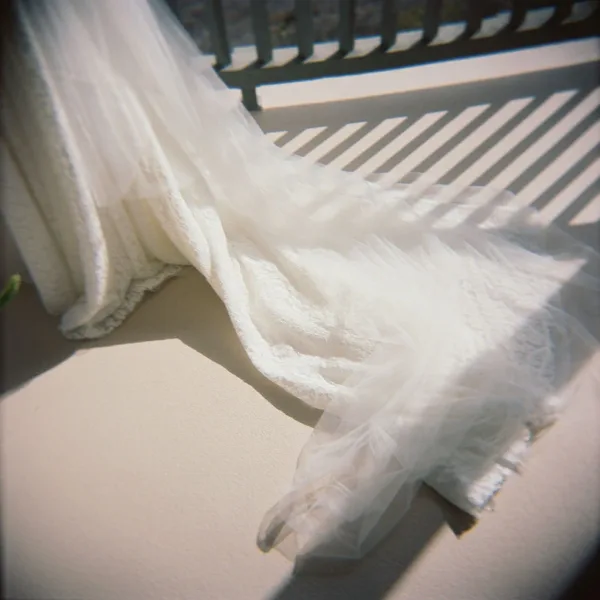 Wedding dress train with lace hem spreading across a sunlit floor, a tulle veil draped over it with balcony railing shadows