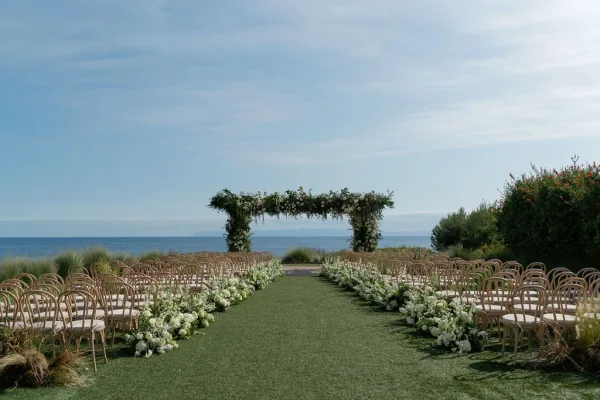 Ceremony setup with an outdoor ceremony aisle lined in white flowers leading to a greenery arch on a lawn overlooking the ocean