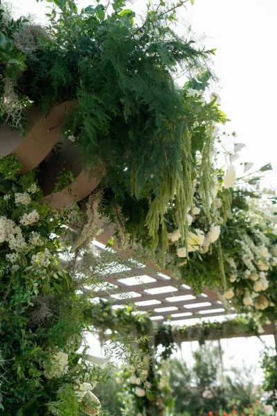Floral ceremony arch with a greenery garland and white flowers, calla lilies, and hanging vines on a pergola in a garden setting
