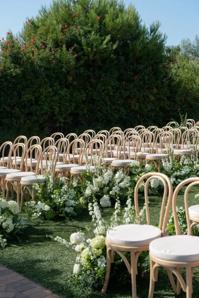 Ceremony aisle decor with white floral arrangements and greenery lining an aisle runner between bentwood chairs on a sunny lawn under blue sky