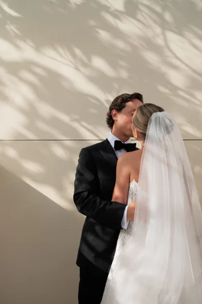 Wedding kiss as bride and groom kissing in a black tuxedo and strapless lace dress, framed by veil and leaf shadows on a beige wall