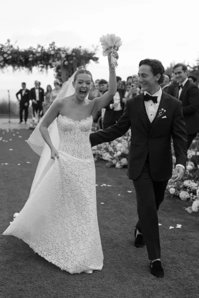 Wedding recessional with bride and groom walking down aisle as she raises her bouquet, guests cheering beneath a floral arch on the lawn