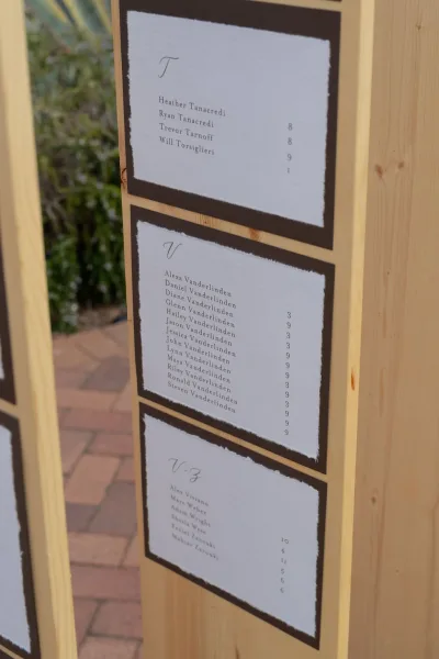 Wedding seating chart escort card display with calligraphy paper cards on black backing, mounted to a wooden board on a brick patio outdoors