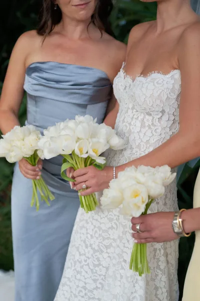 Bridesmaid portrait beside the bride, both holding simple white bouquets, showing veil and lace dress against lush green foliage