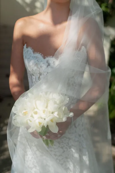 Bridal portrait of a bride in a strapless lace wedding dress holding a white tulip bouquet, veil draped, on sunlit stone steps outdoors