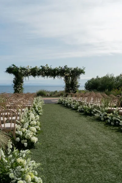 Ceremony setup with a floral arch of white flowers and greenery, wooden chairs and aisle florals on grass, ocean and mountains beyond