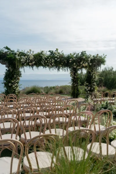 Ceremony setup with a floral arch of white flowers and greenery garland, wood chairs lining an aisle, and an ocean view under cloudy sky