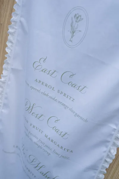 Wedding bar menu with signature cocktails sign in green calligraphy, floral crest and lace-trim napkin styled on woven linen flat lay