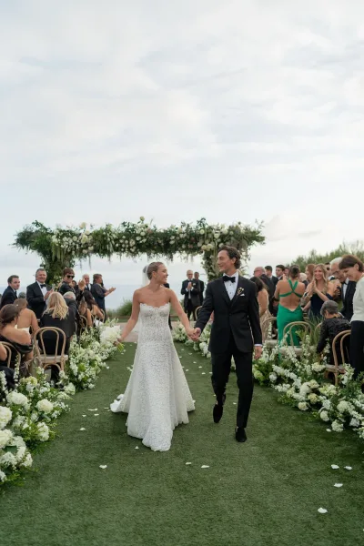 Ceremony recessional as bride and groom walk hand in hand down a floral-lined aisle with white petals, ocean view behind guests