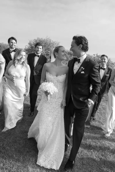 Wedding party portrait of bride and groom walking with bouquets and tuxedos on a grassy lawn with trees under open sky