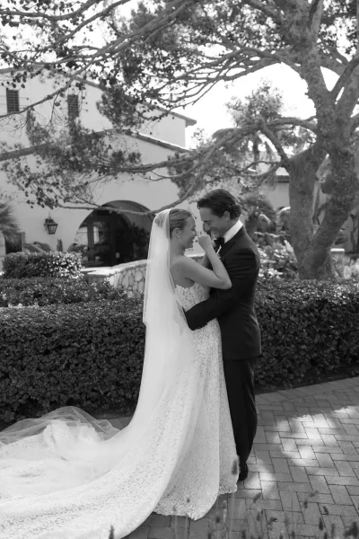Couple portrait in a black and white wedding portrait, bride touching groom’s face as they embrace in a courtyard by an arched doorway and trees