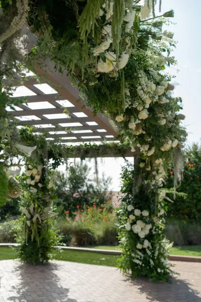 Wedding ceremony arch with white flowers and greenery garland on a pergola, featuring hanging greenery over a garden lawn and brick patio