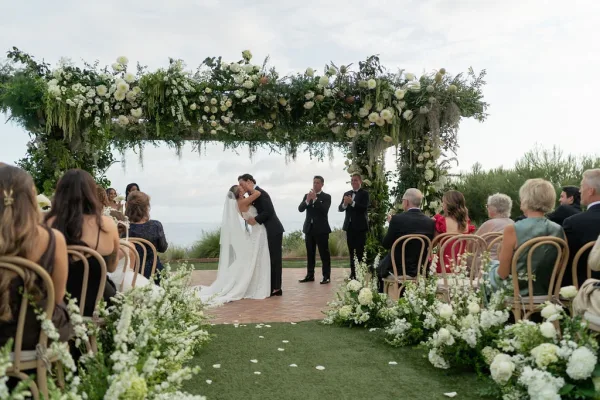 Ceremony kiss as bride and groom embrace beneath a white floral arch with greenery garland, ocean view and cloudy sky behind