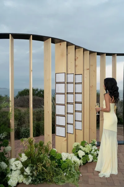 Wedding seating chart on a wood panel display with curved black frame, white hydrangeas at base, on a coastal terrace by the ocean horizon