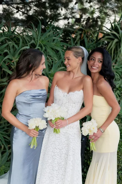 Bride with bridesmaids portrait showing bride in strapless lace gown and veil with friends holding white bouquets against lush tropical greenery