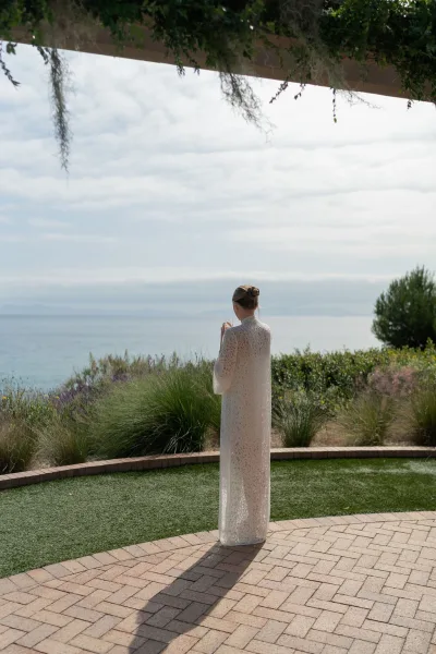 Bridal portrait of bride from behind in a high neck lace wedding dress, holding a champagne flute on a brick patio with ocean view