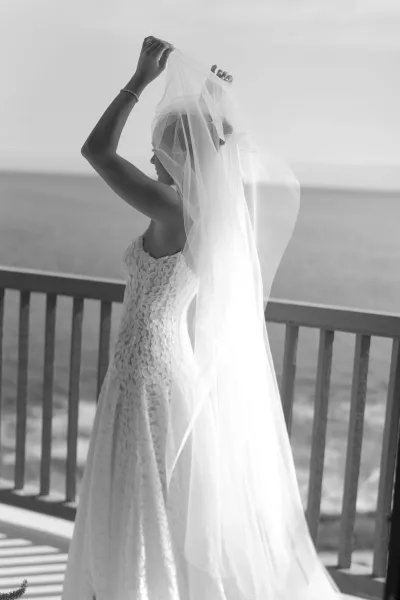Bridal portrait of a bride with veil in side profile, adjusting her lace wedding dress bracelet on a balcony above the ocean and sky