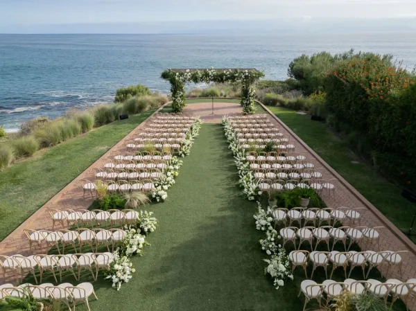 Ceremony setup with oceanfront wedding ceremony aisle, white floral arch and greenery, wooden chairs on a lawn overlooking the coastline