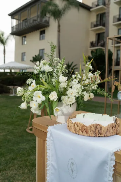 Wedding welcome table decor with white floral arrangement in a ceramic vase, greenery, and escort cards in a basket on a lawn by a tent