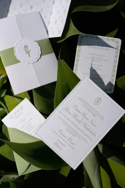 Invitation suite flat lay with green ribbon belly band, embossed monogram seal, and deckled edges on leafy backdrop with sunlight shadows
