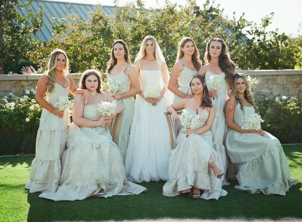 Bridal party portrait with bride and bridesmaids holding white bouquets, bride in veil and pearl necklace on a sunlit garden lawn by a stone wall