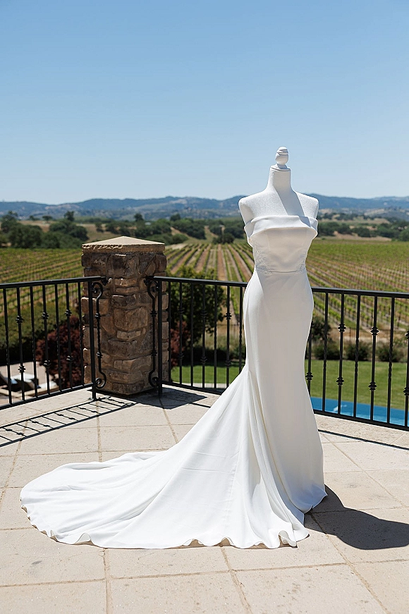 Wedding dress in a strapless wedding dress silhouette on a dress form, chapel train draped on a terrace patio overlooking vineyard hills