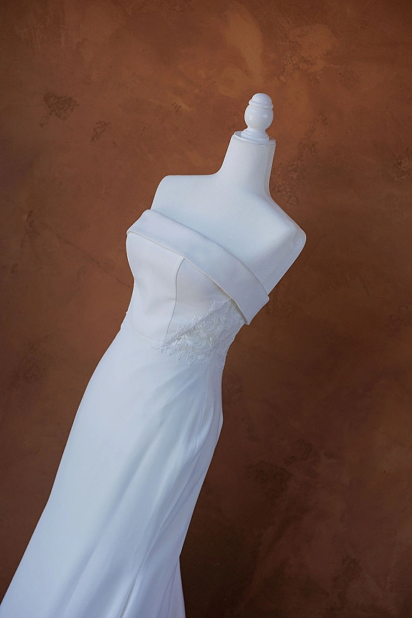 Wedding dress with off the shoulder neckline on a mannequin, featuring lace applique and a fitted silhouette against a warm brown studio backdrop