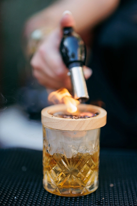 Smoked cocktail with a smoke bubble lid in a whiskey glass over ice, set on a bar mat with a flame against a dark backdrop