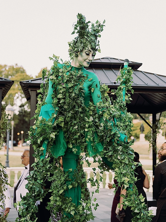 Wedding entertainment stilt walker in a leaf costume with ivy garlands and headpiece, interacting with guests at an outdoor pavilion patio
