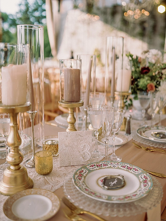 Reception tablescape with wedding table setting of vintage china, lace runner, gold candlesticks and candles under tent string lights
