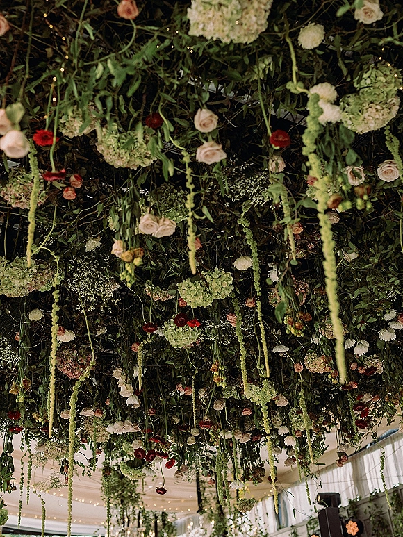 Hanging floral installation with roses, hydrangeas, and cascading amaranthus under a draped tent ceiling with string lights