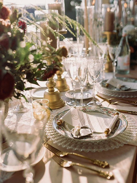Reception tablescape with wedding table place setting, taper candles in brass, floral greenery centerpiece, crystal glasses, gold flatware in sunlight