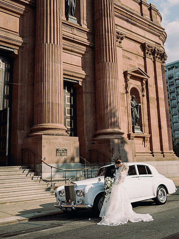 Wedding couple portrait of bride and groom kiss beside a vintage white car on steps of a grand columned building under blue sky
