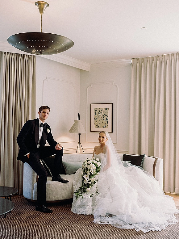 Couple portrait of bride and groom seated on a sofa in a hotel suite, bride in lace dress with veil holding bouquet, groom in tuxedo