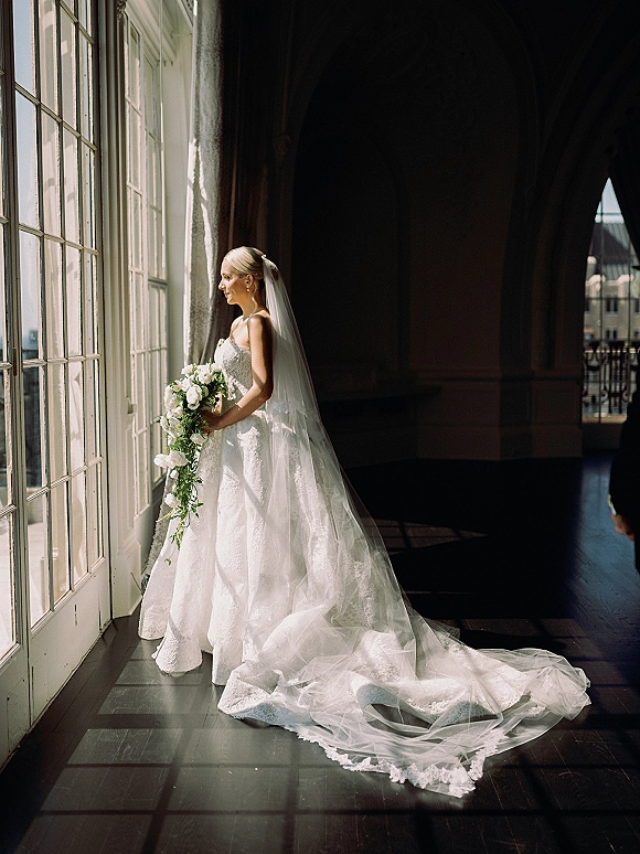 Bridal portrait of a bride by window in a strapless lace gown with long cathedral veil train, holding white flowers in a sunlit hallway