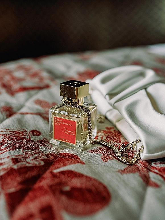 Wedding perfume flatlay with bridal perfume photo styling, featuring a perfume bottle with gold cap and chain necklace on white satin atop red toile bedspread