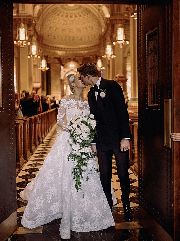 Wedding kiss portrait of bride and groom kissing in a grand church aisle, bride in long-sleeve lace gown holding a cascading bouquet