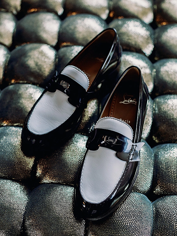Groom wedding shoes, black and white loafers with metal buckles resting on a tufted metallic leather sofa for getting-ready details