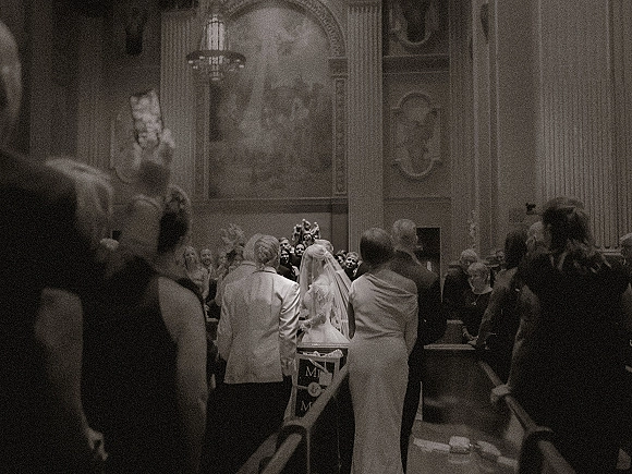 Wedding processional with bride walking down aisle in a long veil and dress, escorted past church pews beneath a chandelier as guests watch