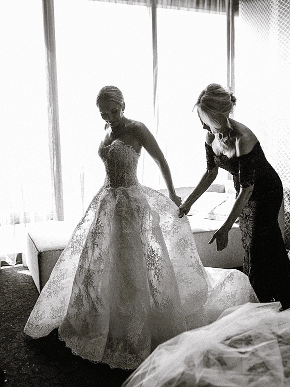 Bride getting ready in a strapless lace wedding dress as her mother helps with final touches in a hotel room by large windows